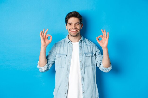 Homem adulto sorridente confiante, mostrando sinais de ok e parecendo satisfeito, como algo, de pé contra um fundo azul.