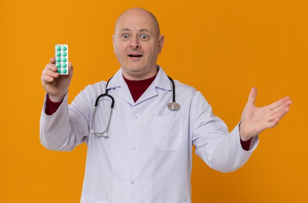 Homem adulto surpreso em uniforme de médico com estetoscópio segurando a embalagem de remédio e mantendo a mão aberta