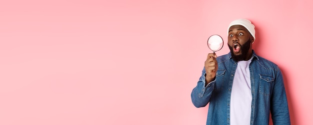 Foto grátis homem africano-americano feliz olhando através da lupa sorrindo espantado em pé contra as costas rosa