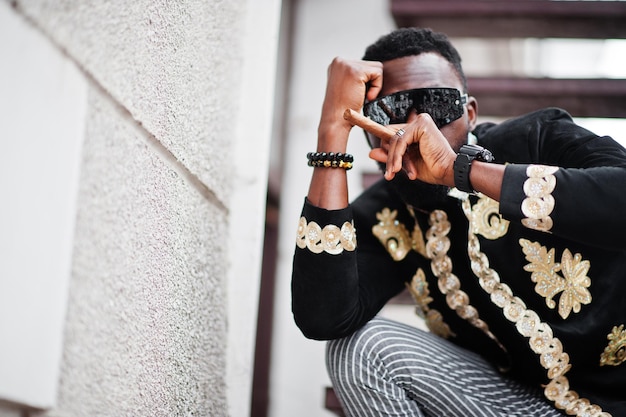 Homem africano mega estiloso em pose de jaqueta tradicional Cara negro na moda em óculos de sol com charuto