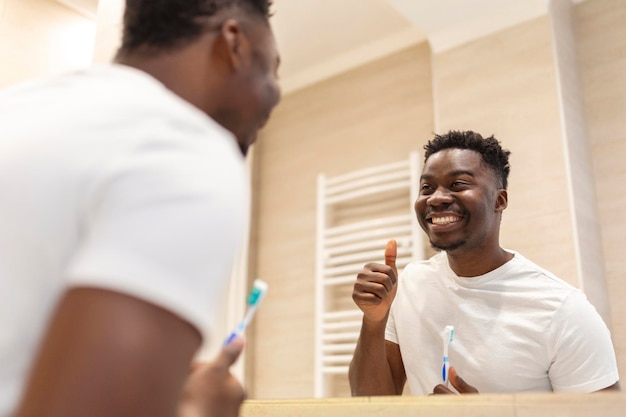 Homem africano sorridente com escova de dentes limpando os dentes e espelho no banheiro