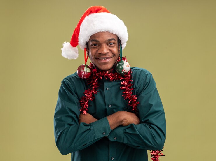Foto grátis homem afro-americano alegre com chapéu de papai noel com guirlanda e bolas de natal nas orelhas se divertindo olhando para a câmera em pé sobre fundo verde.