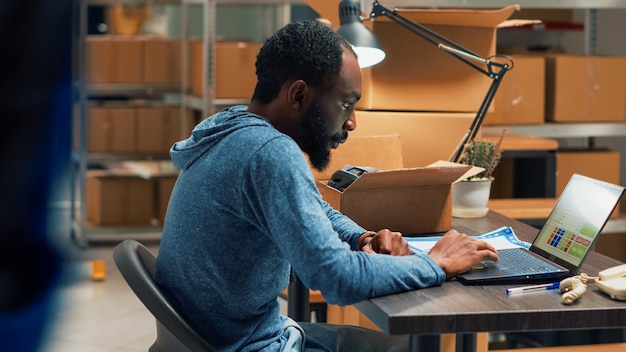 Foto grátis homem afro-americano analisando estoque e pedidos, fazendo inventário no laptop no espaço do armazém. funcionário do sexo masculino usando logística e planejamento financeiro para enviar mercadorias para plano de negócios.