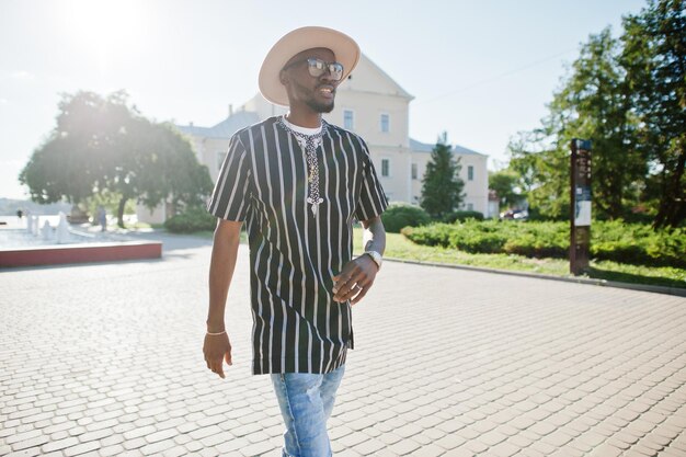 Homem afro-americano atraente com chapéu de camisa listrada e óculos de sol andando na rua no centro da cidade