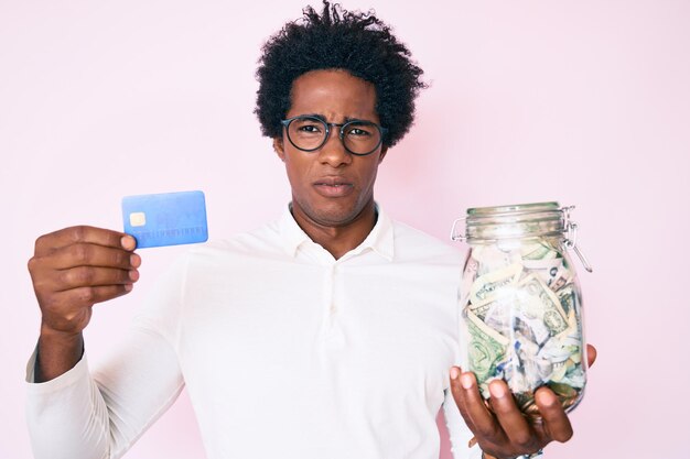 Homem afro-americano bonito com cabelo afro segurando carro de crédito e pote com dólares sem noção e conceito de dúvida de expressão confusa