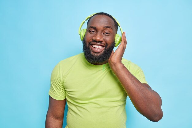 Homem afro-americano bonito e feliz com barba espessa mantém a mão em fones de ouvido estéreo e desfruta de um som perfeito usa uma camiseta verde casual isolada sobre a parede azul