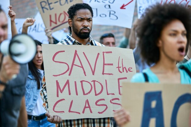 Homem afro-americano carregando faixa com inscrição 'salvar a classe média' enquanto protestava com multidão de pessoas nas ruas da cidade