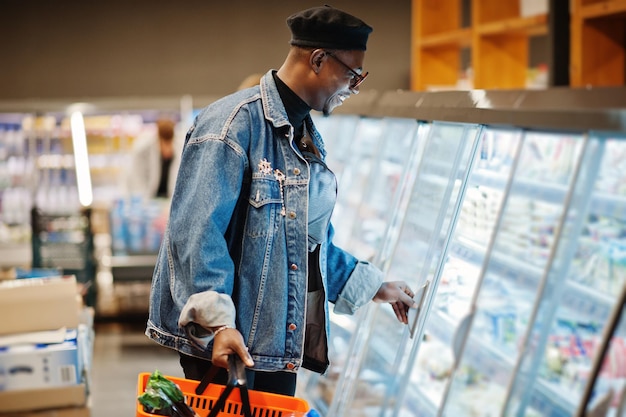 Homem afro-americano casual elegante na jaqueta jeans e boina preta segurando cesta em pé perto de geladeira gastronômica e compras no supermercado