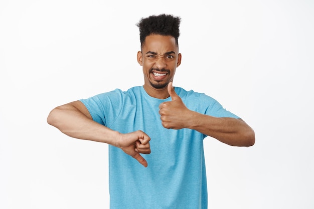 Foto grátis homem afro-americano cético, mostrando os polegares para cima, franzindo a testa e fazendo caretas, inseguro, confuso como avaliar o produto, positivo ou negativo, gosta ou não, fundo branco