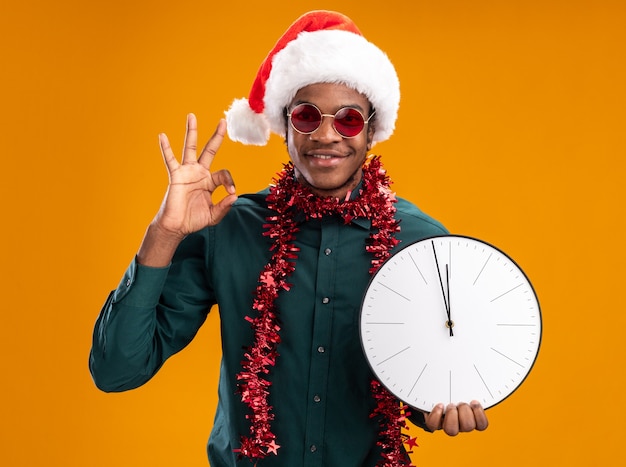 Homem afro-americano com chapéu de papai noel com festão usando óculos escuros, segurando um relógio, olhando para a câmera, sorrindo, mostrando uma placa de ok em pé sobre um fundo laranja