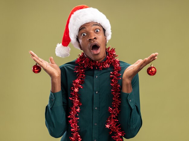 Homem afro-americano com chapéu de Papai Noel com guirlanda segurando bolas de natal, olhando para a câmera espantado e surpreso em pé sobre fundo verde