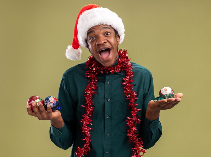 Foto grátis homem afro-americano com chapéu de papai noel com guirlanda segurando bolas de natal, olhando para a câmera, sorrindo com uma carinha feliz em pé sobre um fundo verde