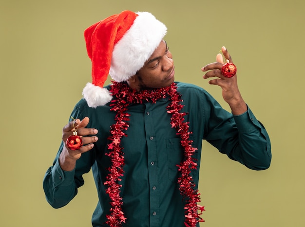 Foto grátis homem afro-americano com chapéu de papai noel com guirlanda segurando bolas de natal, parecendo confuso, tendo dúvidas em pé sobre fundo verde