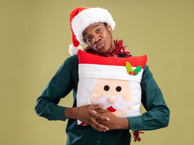 Homem afro-americano com chapéu de Papai Noel com guirlanda segurando travesseiro de natal, olhando para a câmera, feliz e positivo em pé sobre fundo verde