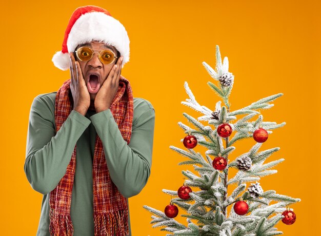 Homem afro-americano com chapéu de Papai Noel e lenço ao redor do pescoço espantado e surpreso ao lado de uma árvore de Natal sobre uma parede laranja