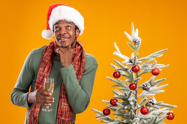 Homem afro-americano com chapéu de Papai Noel e lenço ao redor do pescoço segurando uma taça de champanhe, olhando perplexo ao lado da árvore de Natal sobre fundo laranja