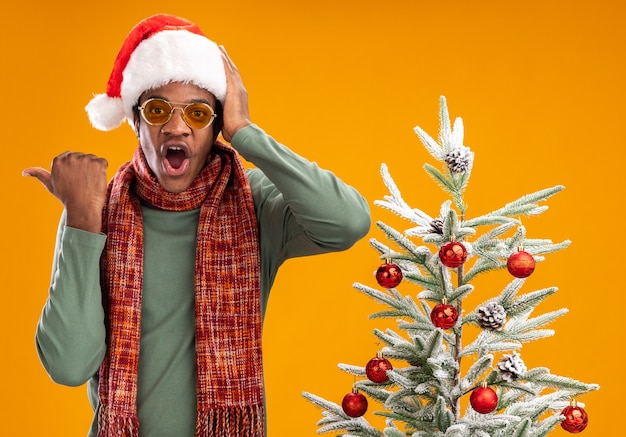 Homem afro-americano com chapéu de papai noel e lenço em volta do pescoço olhando para a câmera maravilhado, apontando com o polegar para o lado, ao lado de uma árvore de natal sobre fundo laranja