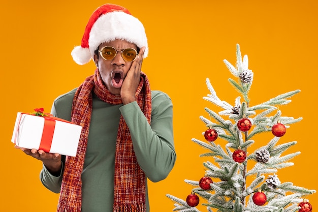 Homem afro-americano com chapéu de Papai Noel e lenço em volta do pescoço segurando um presente, olhando para a câmera espantado e surpreso ao lado de uma árvore de Natal sobre fundo laranja