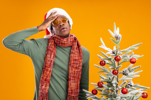 Homem afro-americano com chapéu de papai noel e lenço no pescoço, cansado e entediado, em pé ao lado de uma árvore de natal sobre uma parede laranja