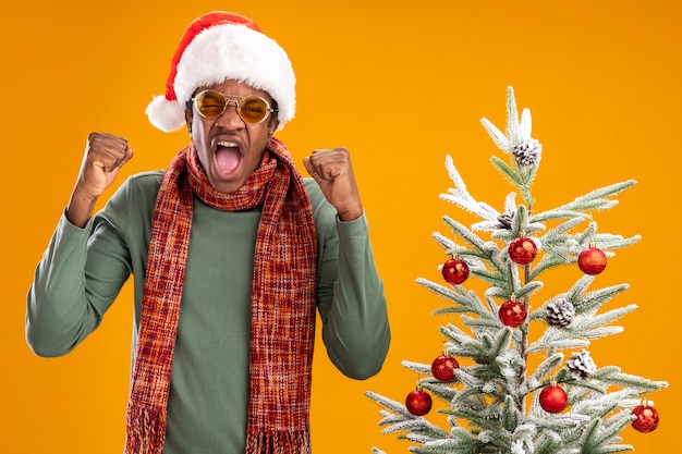 Foto grátis homem afro-americano com chapéu de papai noel e lenço no pescoço cerrando os punhos zangado, louco, louco, ao lado de uma árvore de natal sobre fundo laranja