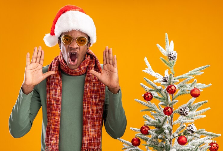 Foto grátis homem afro-americano com chapéu de papai noel e lenço no pescoço gritando com expressão agressiva ao lado de uma árvore de natal sobre um muro de laranja