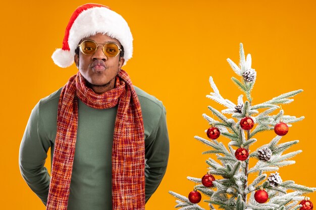 Homem afro-americano com chapéu de Papai Noel e lenço no pescoço, olhando para a câmera, mantendo os lábios como se fosse beijar em pé ao lado de uma árvore de natal sobre fundo laranja