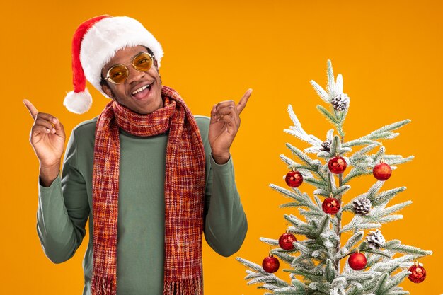 Homem afro-americano com chapéu de Papai Noel e lenço no pescoço sorrindo alegremente ao lado de uma árvore de Natal sobre um muro laranja