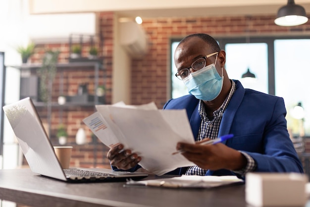 Homem afro-americano com máscara facial, analisando documentos para planejar a estratégia de marketing. Funcionário da empresa trabalhando com laptop e cuidando da papelada do projeto durante a pandemia de coronavírus.