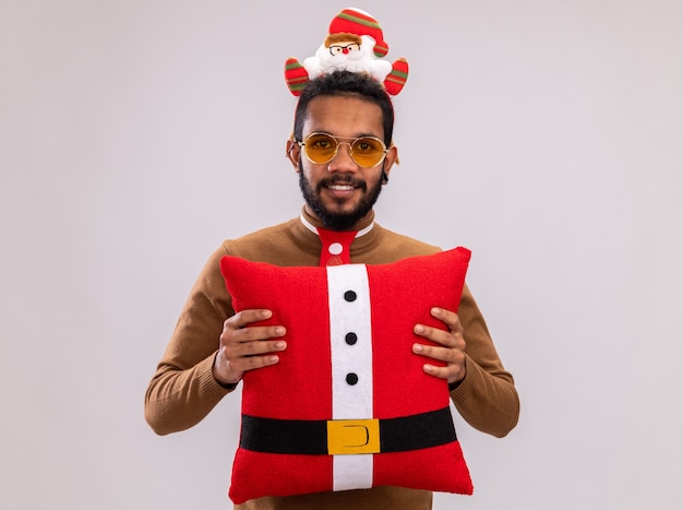 Foto grátis homem afro-americano com suéter marrom e papai noel na cabeça com gravata vermelha engraçada segurando a almofada de natal, olhando para a câmera com um sorriso no rosto em pé sobre um fundo branco