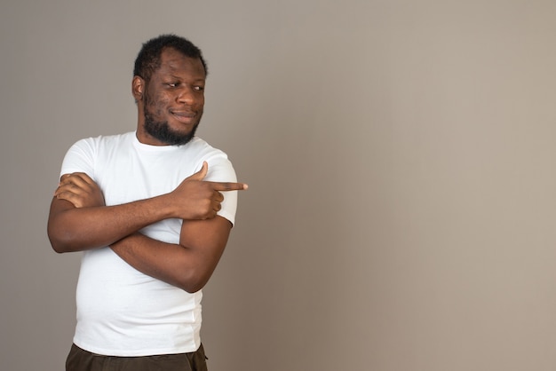 Foto grátis homem afro-americano de braços cruzados, apontando para a esquerda com uma das mãos, em frente à parede cinza.