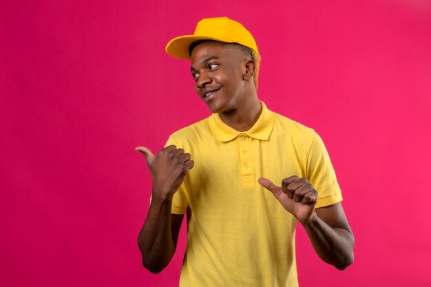 Homem afro-americano de entrega em camisa pólo amarela e boné olhando para o lado apontando para o lado com polegares sorrindo amigável em pé na rosa