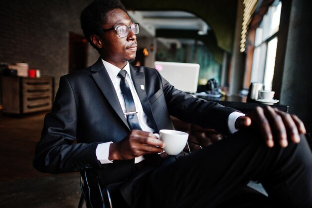 Homem afro-americano de negócios usa terno preto e óculos no escritório bebendo café da manhã antes do dia de trabalho