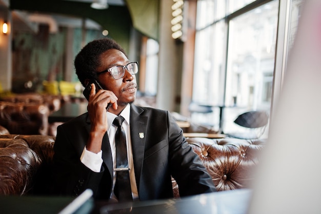 Homem afro-americano de negócios usa terno preto e óculos sentado no escritório e falando no telefone atrás do laptop