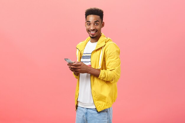 Homem afro-americano elegante encantado com corte de cabelo afro encaracolado em pé meio virado na parede rosa segurando o smartphone vestindo uma jaqueta amarela da moda, sorrindo alegremente, mostrando as características do dispositivo de amigo.