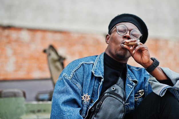 Homem afro-americano em boina de jaqueta jeans e óculos fumando charuto e posou contra veículo blindado militar btr