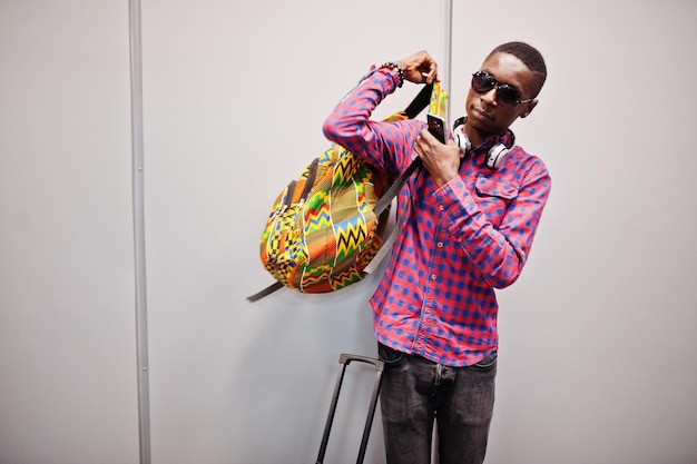 Foto grátis homem afro-americano em óculos escuros de camisa quadriculada e jeans com mala e mochila viajante negro contra parede cinza