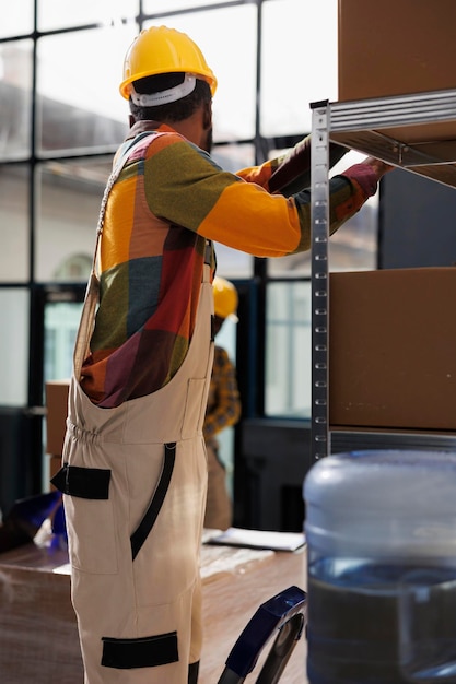 Foto grátis homem afro-americano em pé na escada e procurando encomendas na prateleira em armazém industrial. trabalhador de depósito usando capacete levando caixa de papelão para embalagem de pedidos de clientes no depósito