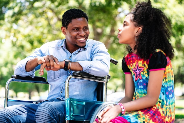 Homem afro-americano em uma cadeira de rodas, curtindo e se divertindo com a filha no parque.