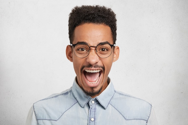 Homem afro-americano emocional com óculos redondos, abre a boca de empolgação e exclama com alegria