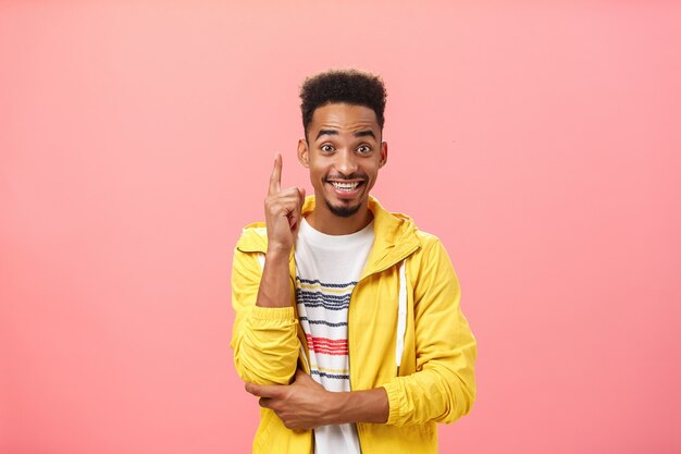 Homem afro-americano entusiasmado e satisfeito, adicionando a sugestão, levantando o dedo indicador em um gesto de eureka e sorrindo alegremente enquanto discute uma invenção ou teoria interessante, sorrindo amplamente sobre a parede rosa