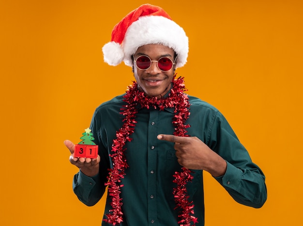 Homem afro-americano feliz e positivo com chapéu de Papai Noel com festão usando óculos escuros segurando cubos de brinquedo com a data de ano novo apontando com o dedo indicador em pé sobre fundo laranja