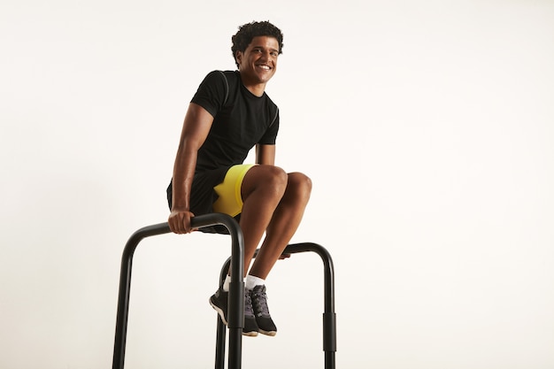 Homem afro-americano feliz e sorridente com roupa de treino sintética preta, fazendo exercícios em casa nas barras paralelas, isolado no branco