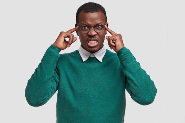 Homem afro-americano frustrado e insatisfeito mantém os dois dedos indicadores nas têmporas, tem uma expressão facial irritada e insatisfeita, franze a testa com raiva