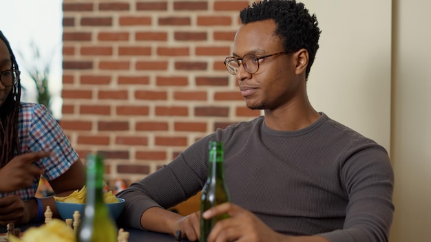 Homem afro-americano jogando xadrez com amigos, servindo bebidas alcoólicas e lanches no apartamento. grupo de pessoas felizes se divertindo se reunindo com cerveja e jogos de tabuleiro.