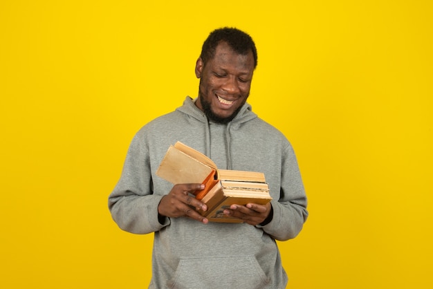 Homem afro-americano lendo livros na mão, em pé sobre a parede amarela.