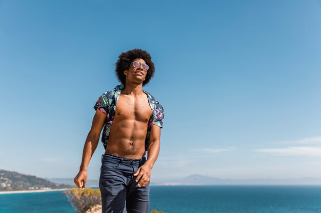 Homem afro-americano muscular considerável que levanta no fundo azul do céu e do mar