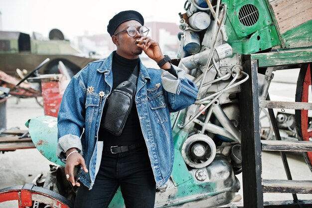 Homem afro-americano na boina de jaqueta jeans e óculos fumando charuto e posou contra o antigo veículo retrô