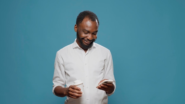 Homem afro-americano navegando na internet no smartphone e segurando a xícara de café na mão, aproveitando o intervalo do trabalho corporativo. Jovem mandando mensagens online no celular para relaxar.