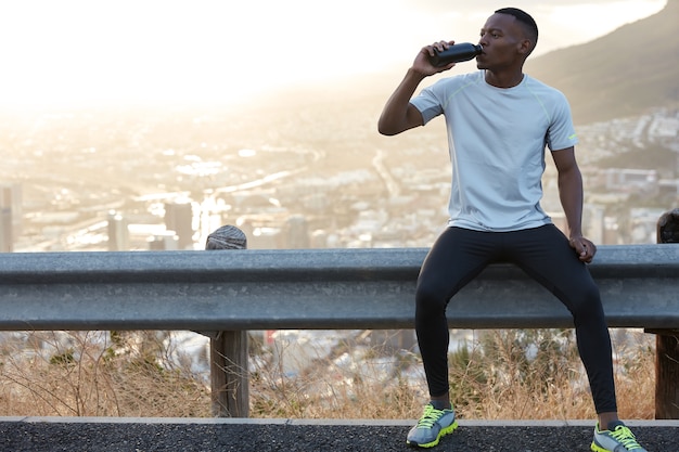 Homem afro-americano sedento bebe água doce, aproveita o intervalo após os treinamentos esportivos ao ar livre, senta-se em uma placa de estrada com uma cópia da vista panorâmica da montanha para conteúdo promocional ou informações