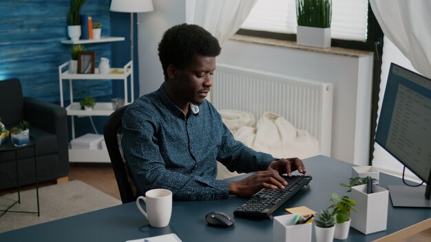 Homem afro-americano sorridente autêntico, trabalhando em casa, usuário de computador de trabalho remoto. Foto em câmera lenta de freelancer usando comunicação on-line com internet na web em apartamento moderno